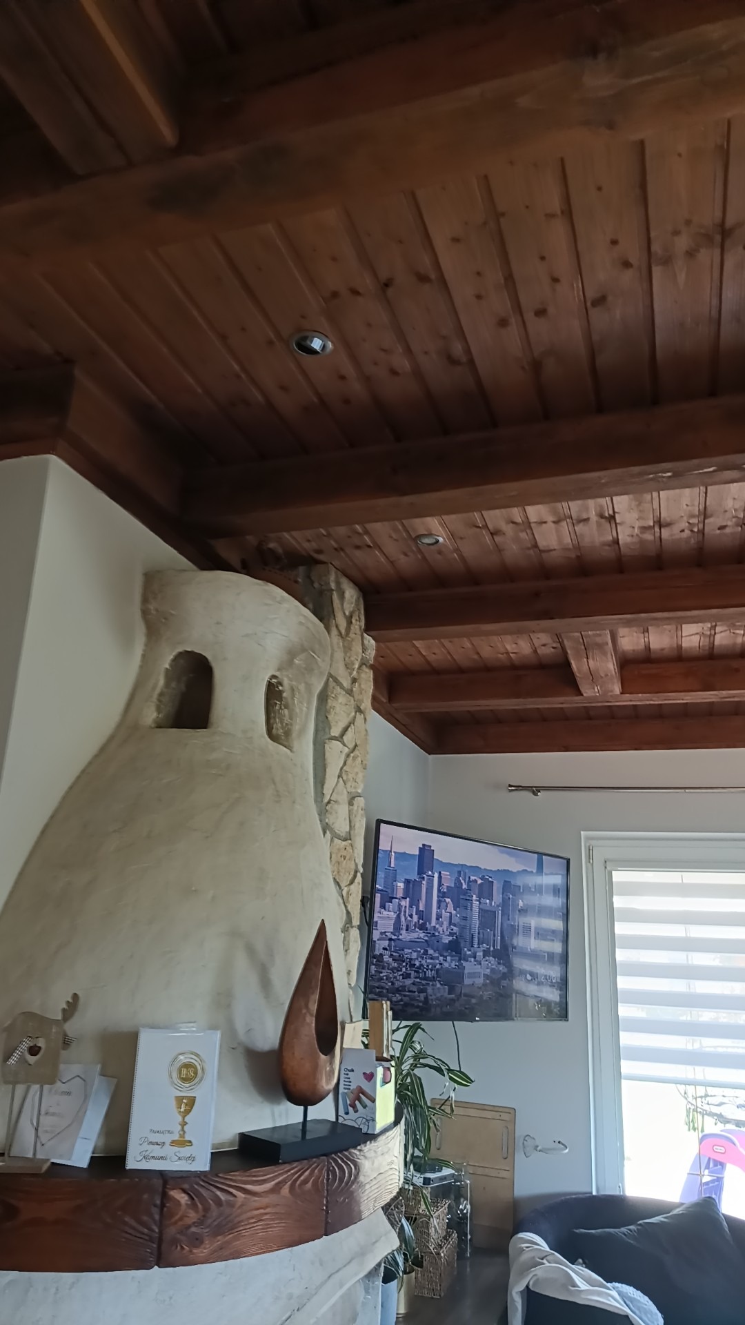 Drewniany sufit z belkami i wbudowanym oświetleniem nad kominkiem z dekoracyjnym tynkiem i kamiennym wykończeniem, obok telewizor z widokiem na miasto.