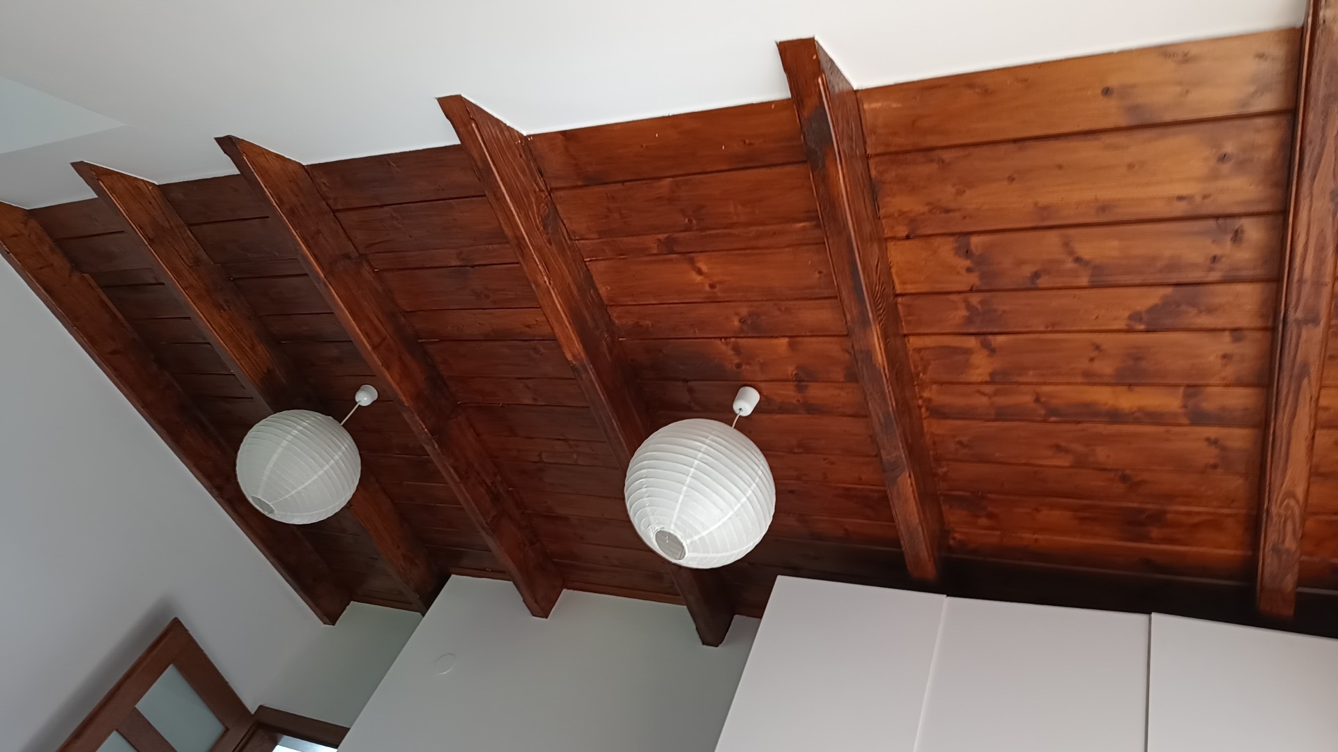 Drewniany sufit z widocznymi belkami i dwoma białymi, okrągłymi lampami papierowymi, widok od dołu.