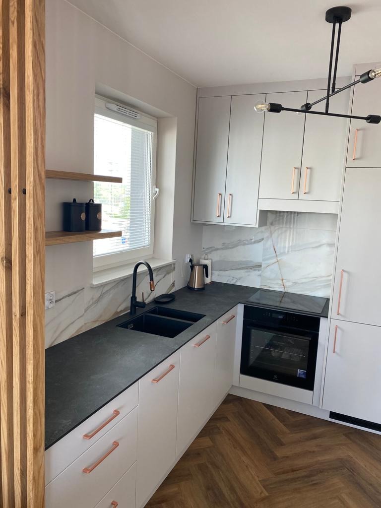 Nowoczesna kuchnia w Myślenicach z białymi szafkami, czarnym blatem i marmurowym backsplashiem. Uwagę zwracają miedziane uchwyty i armatura, oraz parkiet ułożony w jodełkę.