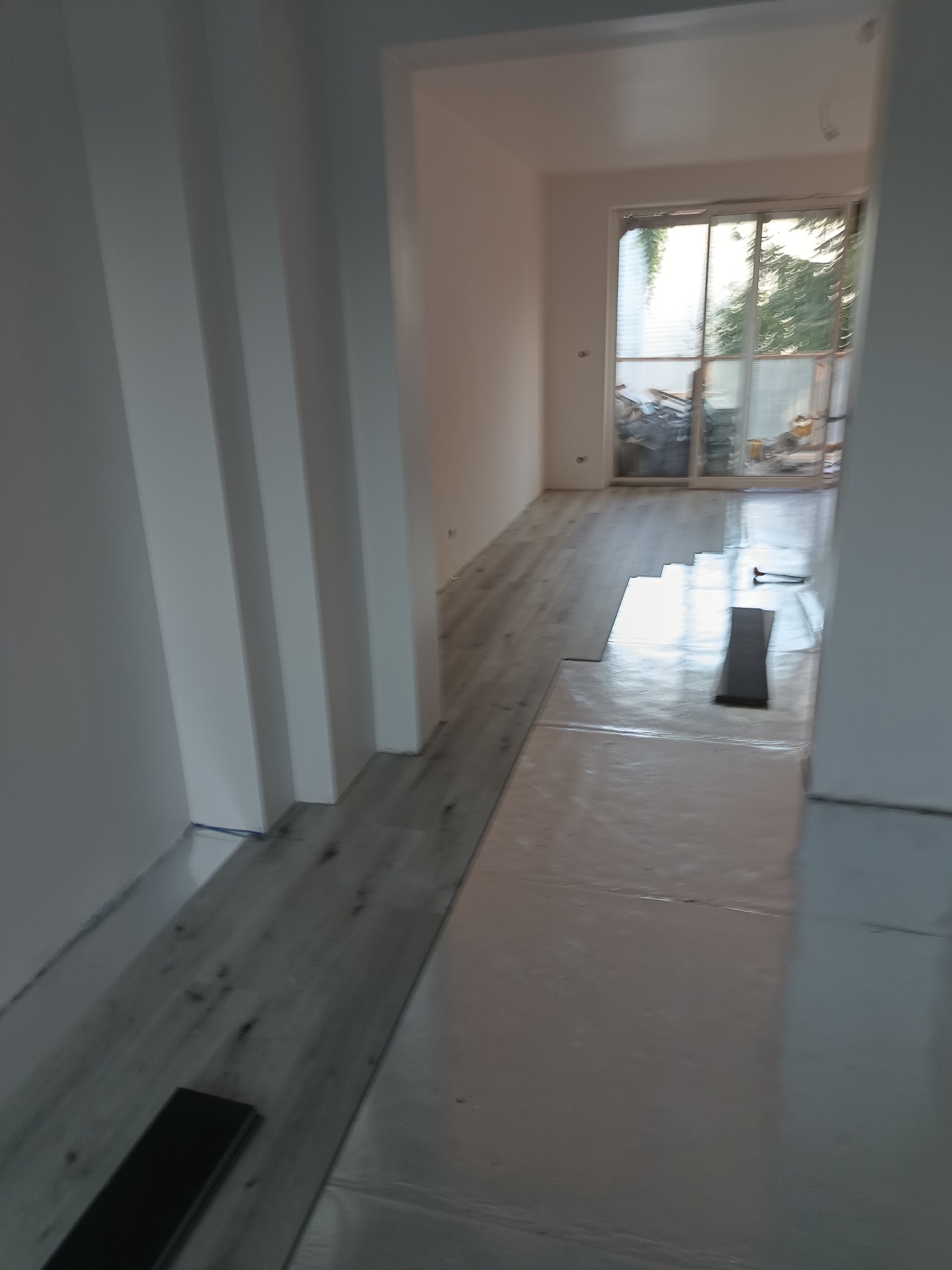 Układanie paneli podłogowych w jasnym, odnawianym wnętrzu z widocznym fragmentem okna balkonowego i przygotowanymi panelami.