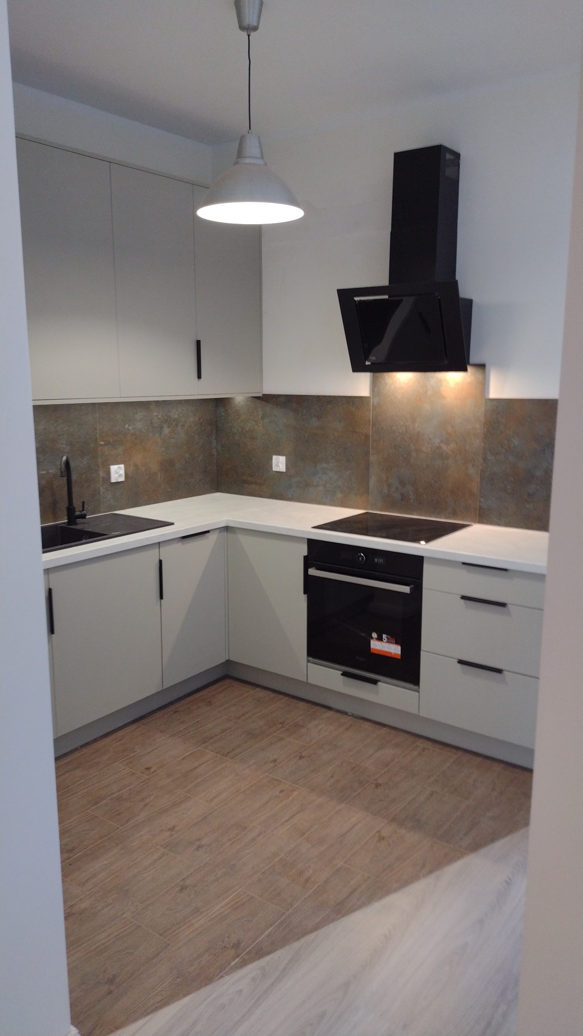 Nowoczesna kuchnia z szarymi szafkami, czarnym okapem i płytą indukcyjną, brązowe płytki imitujące kamień na ścianie, drewnopodobna podłoga z płytek.