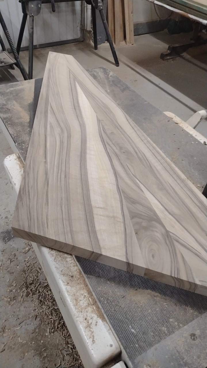 Surowy element drewniany z wyraźnym usłojeniem, leżący na stole roboczym w warsztacie, widoczne ślady obróbki drewna.