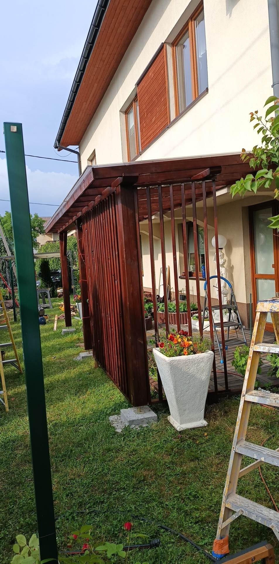 Ciemnobrązowa drewniana pergola z pionowymi listwami, częściowo zasłaniająca taras, z donicą kwiatów i drabiną w ogrodzie przed domem.