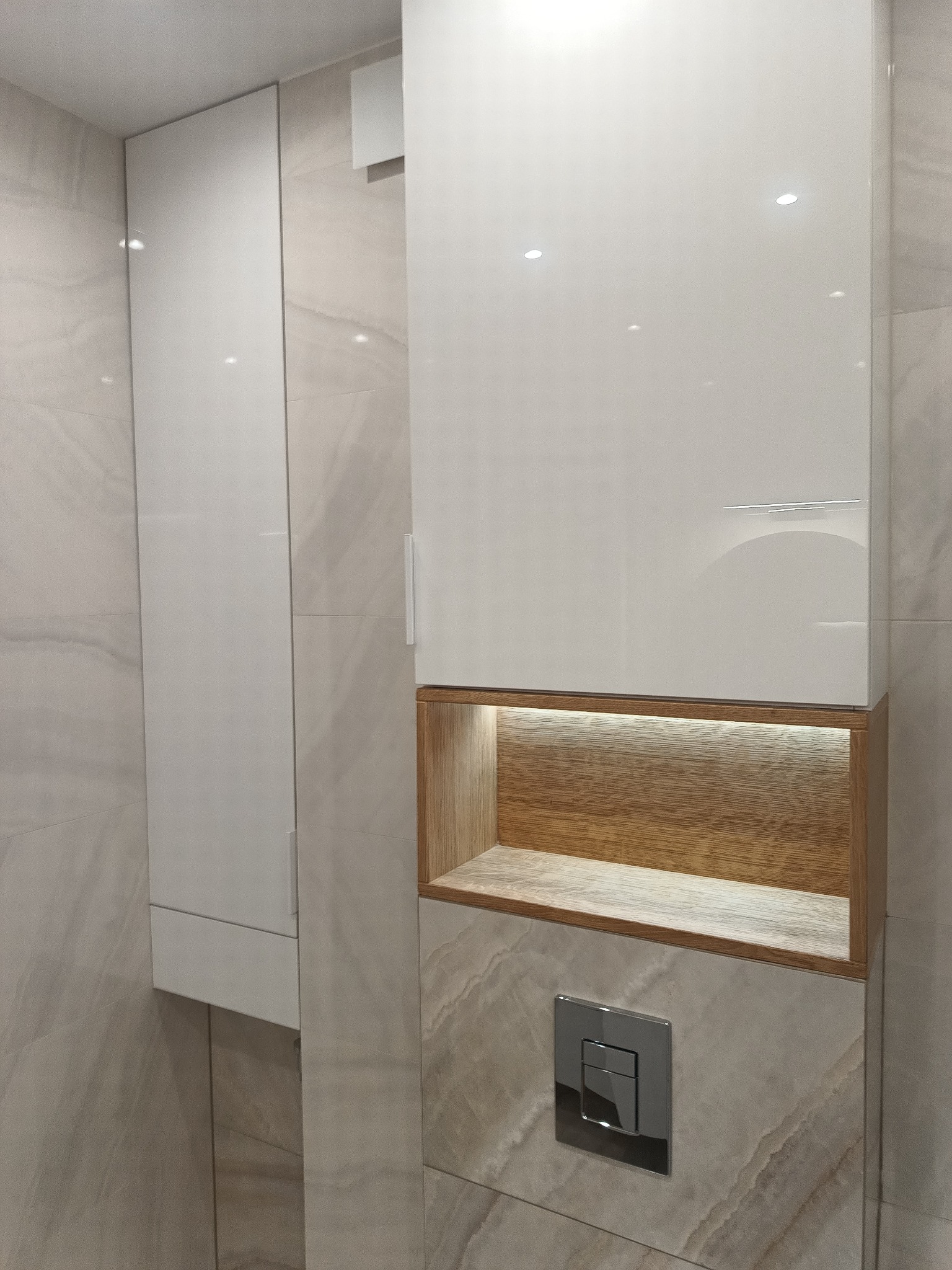Białe, lakierowane szafki łazienkowe z drewnianą wnęką oświetloną LED, zamontowane na ścianie wyłożonej płytkami imitującymi marmur, widoczny przycisk spłuczki.