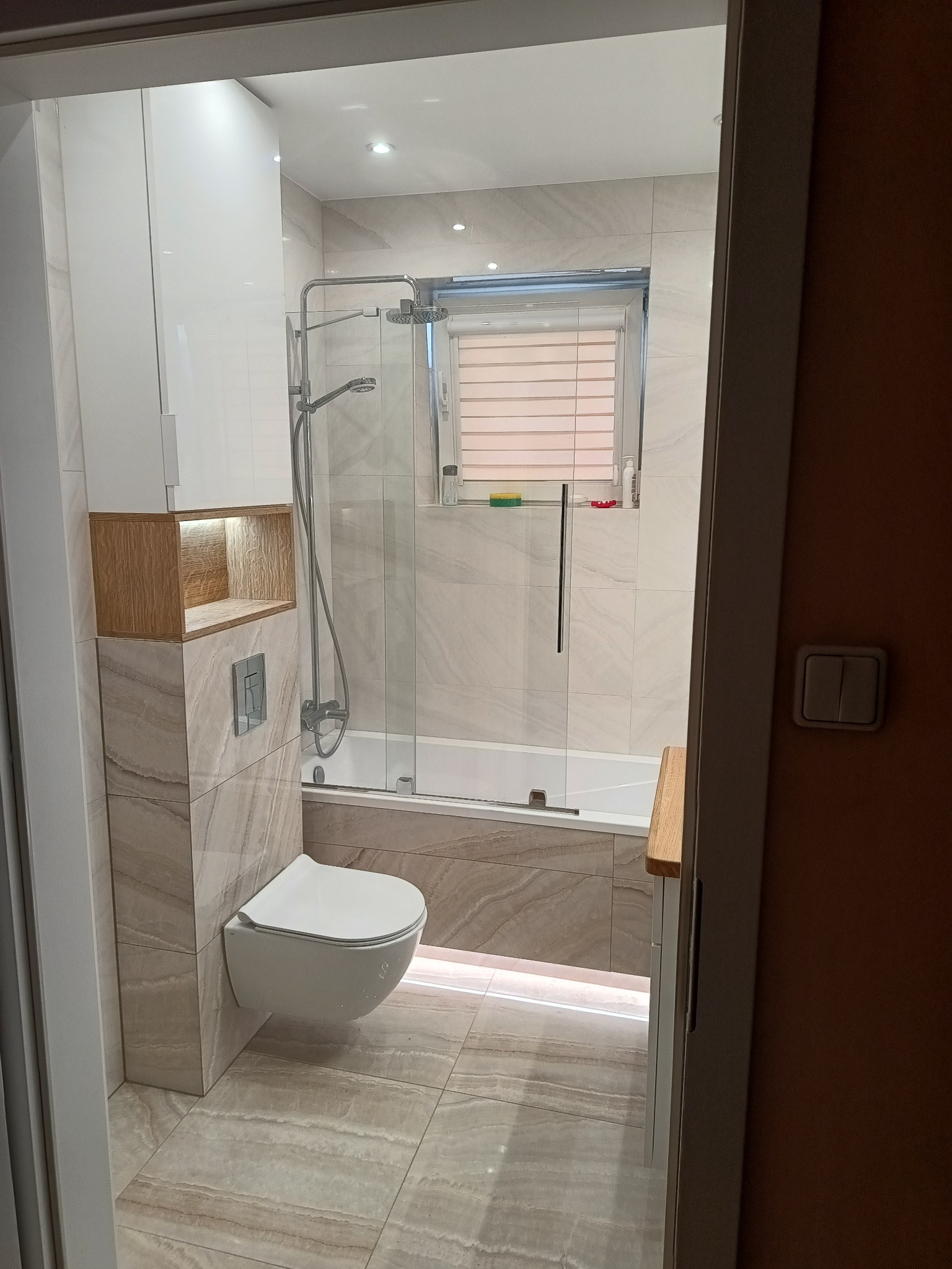 Wykończona łazienka z beżowymi płytkami imitującymi marmur, podwieszana toaleta, zabudowana wanna z parawanem, oświetlenie LED pod wanną i w szafce wnękowej.