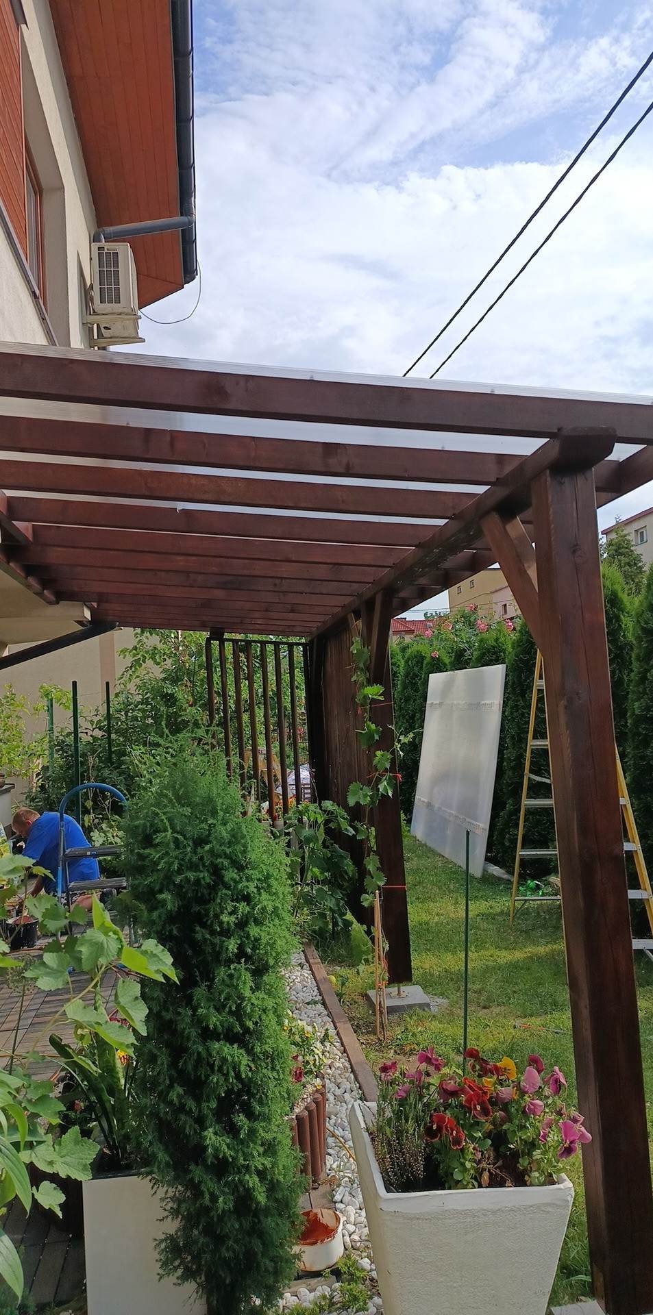 Drewniana pergola z przezroczystym zadaszeniem w przydomowym ogrodzie, widoczne rośliny, drabina i fragment budynku z klimatyzatorem.
