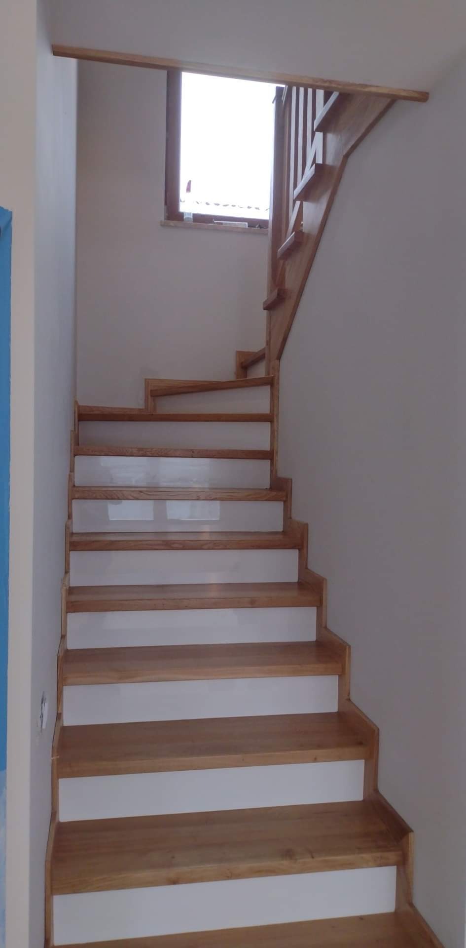 Drewniane schody z białymi podstopnicami, widok z dołu, naturalne oświetlenie z okna na półpiętrze.