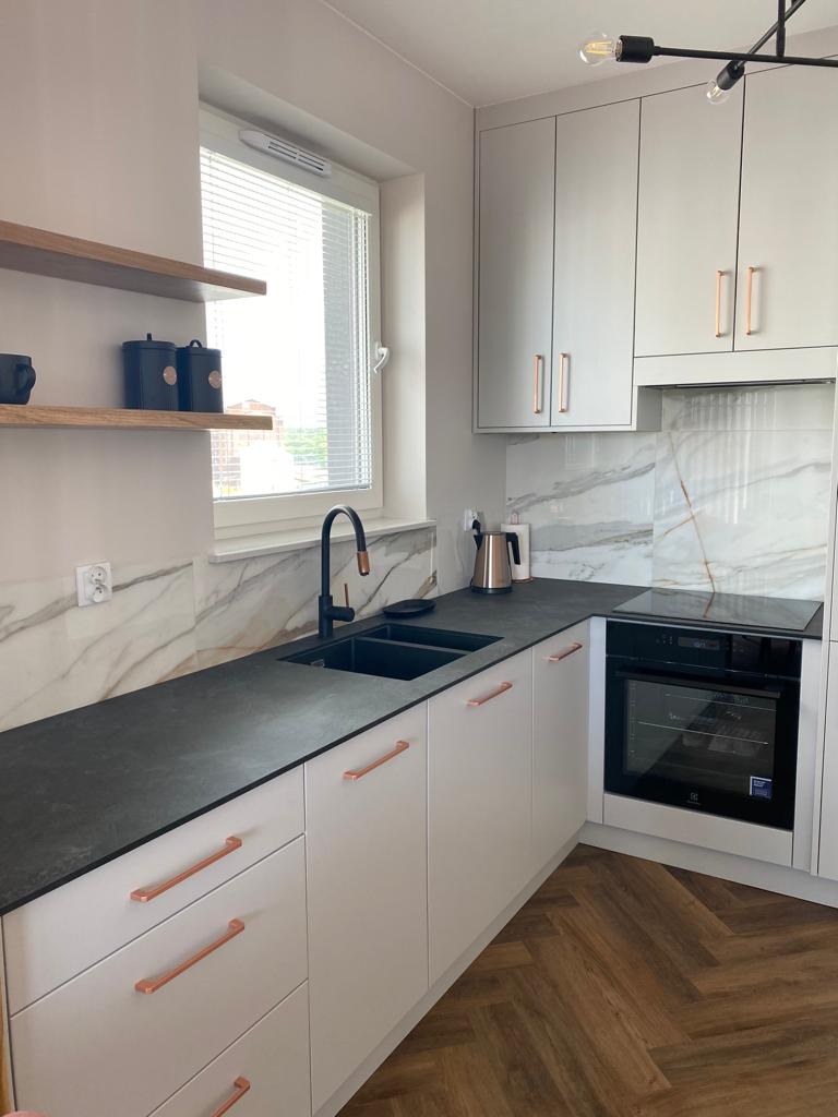 Nowoczesna kuchnia z białymi szafkami, czarnym blatem i miedzianymi uchwytami, marmurowy backsplash, drewniana podłoga w jodełkę.