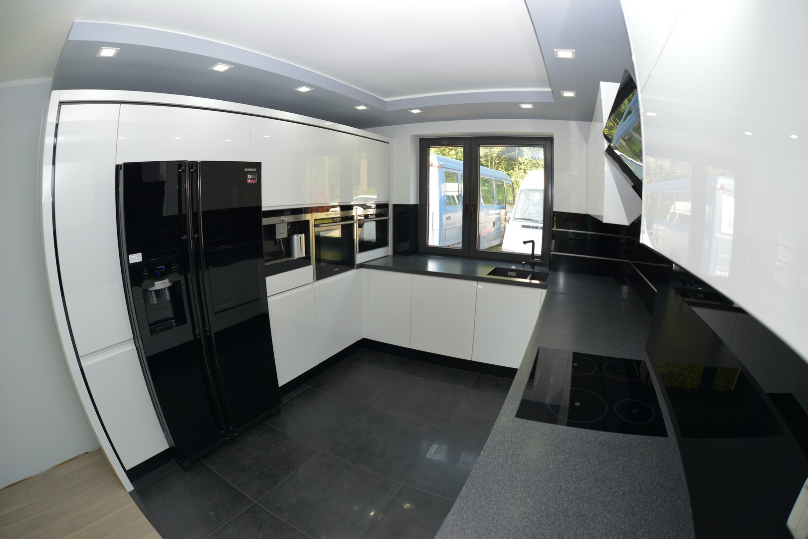 Nowoczesna kuchnia z białymi lakierowanymi szafkami i czarnymi blatami, wyposażona w czarną lodówkę side-by-side i płytę indukcyjną, widok z szerokiego kąta.