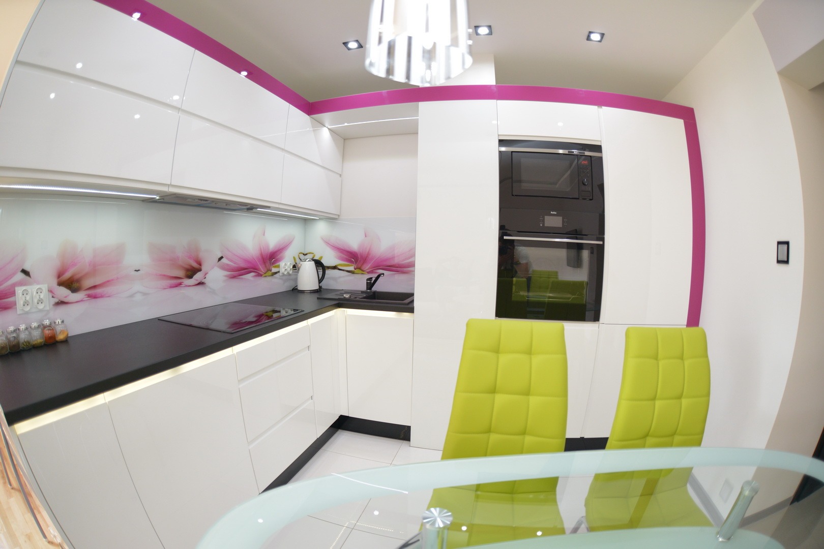 Biała kuchnia z frontami na wysoki połysk, czarnym blatem i panelem nad blatem z motywem magnolii, akcentowana różowymi listwami. Widoczne wbudowane sprzęty AGD i krzesła w kolorze limonkowym.