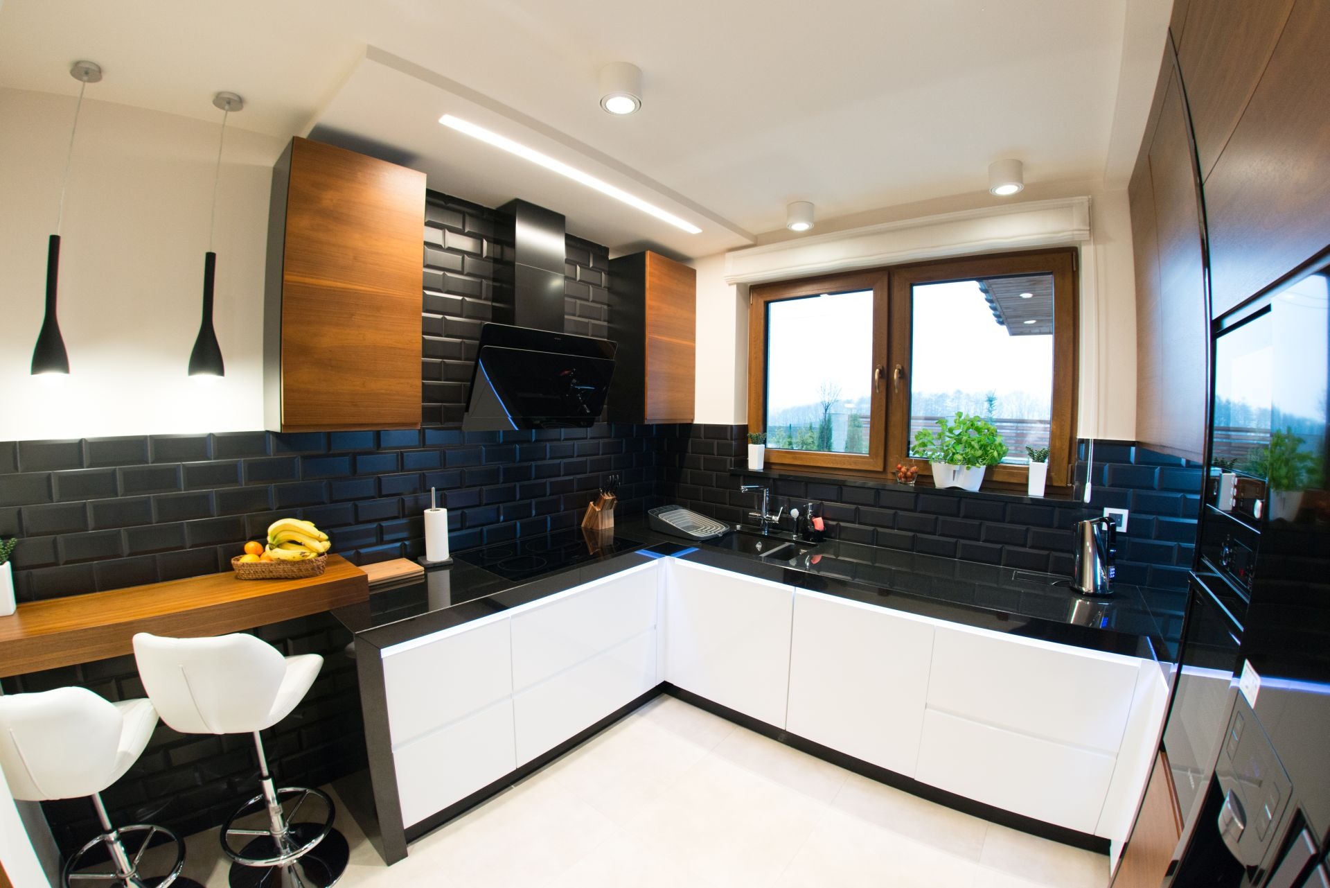 Nowoczesna kuchnia z czarnymi płytkami metra, białymi szafkami z podświetleniem LED, drewnianymi akcentami i oknem z widokiem na zieleń.