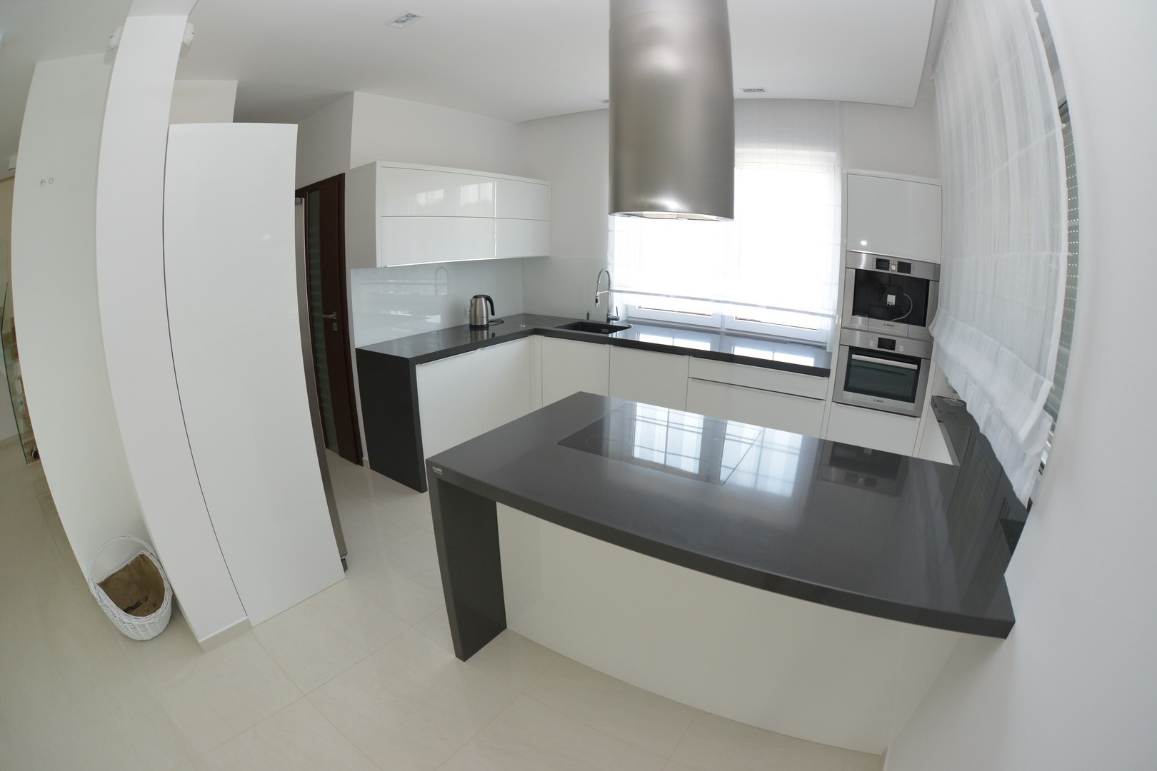 Nowoczesna, minimalistyczna kuchnia z białymi szafkami i ciemnoszarym blatem, widok z szerokiego kąta, srebrny okap nad wyspą kuchenną.