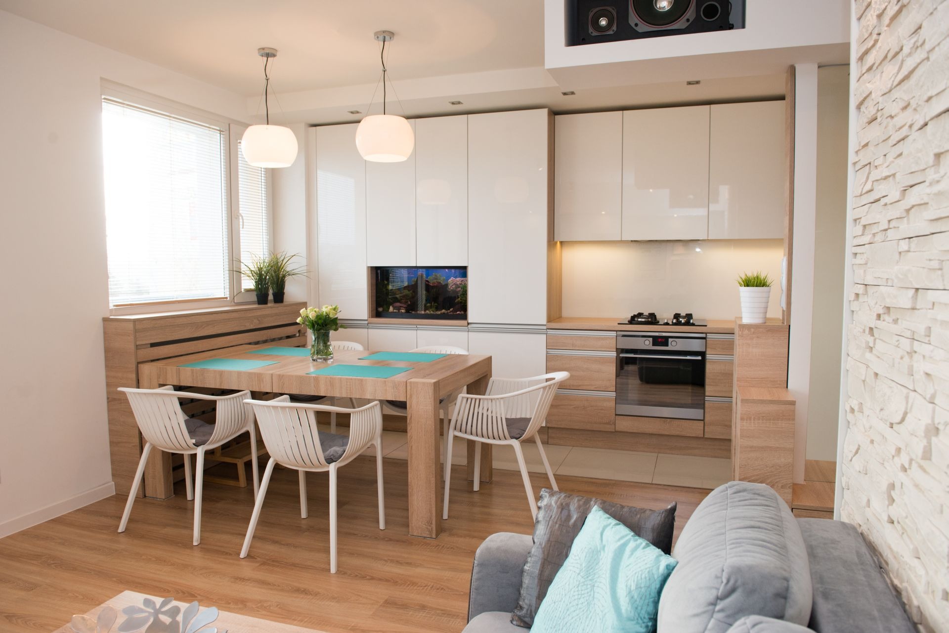 Jasna kuchnia z białymi frontami i drewnianymi akcentami, z wbudowanym akwarium, widok na stół jadalny z czterema krzesłami i część salonu z szarą sofą i turkusową poduszką.