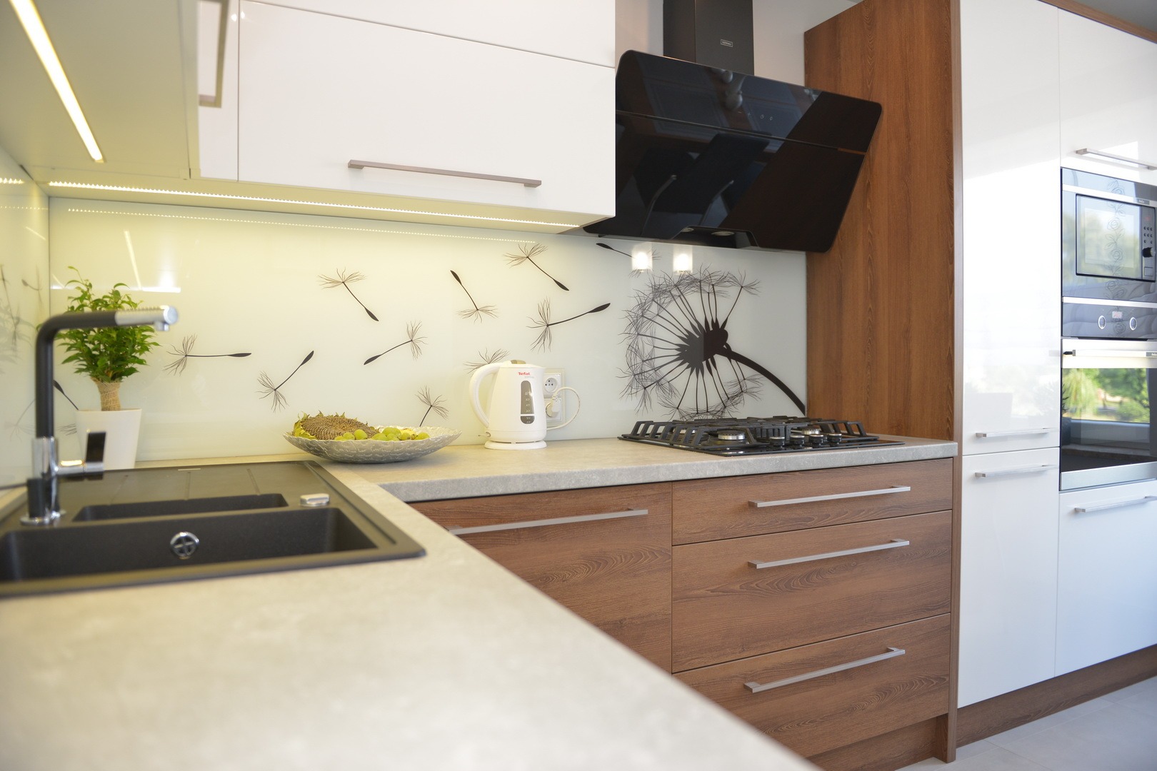 Nowoczesna kuchnia z białymi i drewnopodobnymi frontami, szklanym panelem z motywem dmuchawca, czarnym okapem i szarą armaturą. Na blacie czajnik elektryczny i misa z owocami.