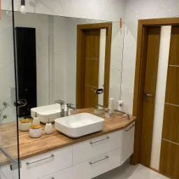 Nowoczesna łazienka z białymi płytkami imitującymi marmur, drewnianym blatem pod umywalką i białymi szafkami z szufladami oraz dużym lustrem.