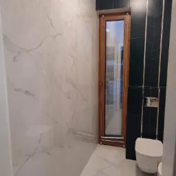 Nowoczesna łazienka z białymi marmurkowymi płytkami na ścianach i podłodze, kontrastującymi z ciemnymi płytkami ułożonymi w jodełkę wokół drewnianego okna i podwieszaną toaletą.