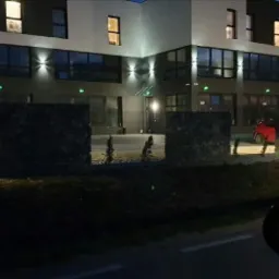 Nowoczesny budynek biurowy oświetlony zewnętrznymi kinkietami LED, widok z ulicy w nocy, z widocznym fragmentem lusterka samochodowego w prawym dolnym rogu kadru.