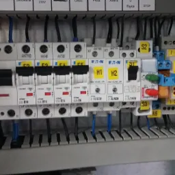 Rozdzielnica elektryczna z wyłącznikami Eaton, bezpiecznikami i elementami sterującymi, widoczne przewody i oznaczenia, zamontowana w szarej obudowie.