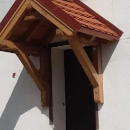 Drewniany daszek nad wejściem z czerwoną dachówką, wsparty na solidnych, drewnianych wspornikach, zamontowany nad ciemnymi drzwiami wejściowymi do budynku o białej, chropowatej elewacji.