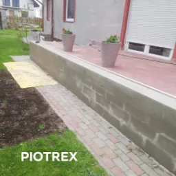 Świeżo wymurowany murek z betonowych bloczków, z wykończonym cokołem, obok ułożona ścieżka z czerwono-szarej kostki, w tle dom z roletami i donice z kwiatami.