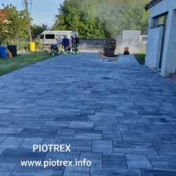 Ekipa montuje szarą kostkę brukową na podjeździe przed domem, zagęszczarka w użyciu, widoczny firmowy bus i logo firmy na kostce.