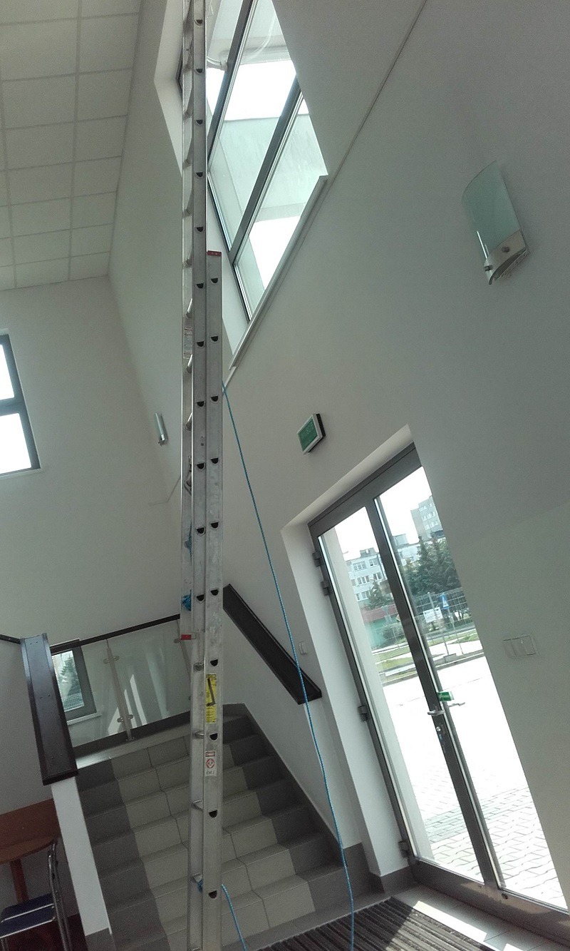 Wysoka, rozłożona drabina aluminiowa oparta o ścianę z oknami na wysokości wewnątrz jasnego budynku, niebieska lina zabezpieczająca, widok z dołu w górę.