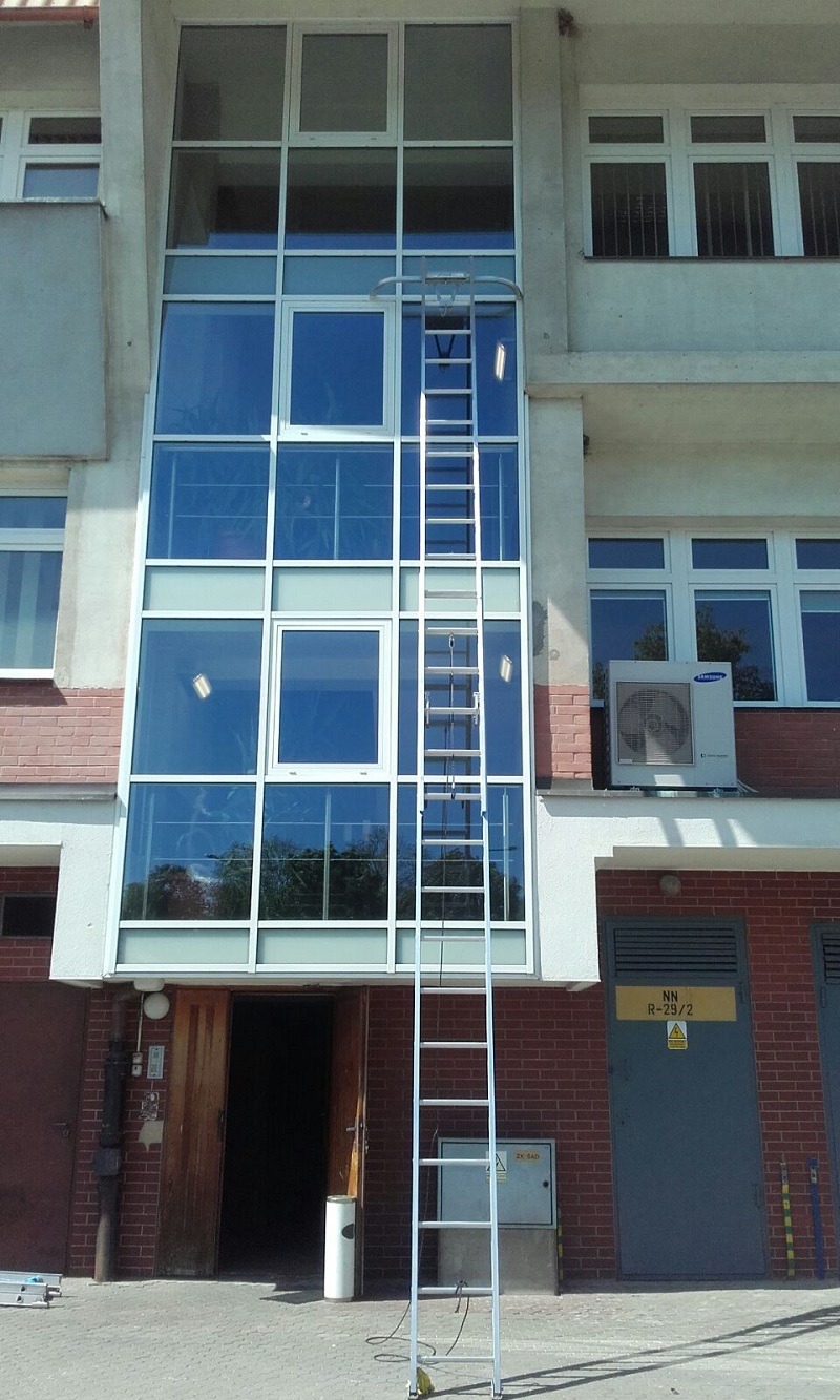 Wysoka ściana przeszklona z wieloma oknami, do których dostawiona jest długa, aluminiowa drabina z zabezpieczeniem górnym, umieszczona na chodniku przed budynkiem.