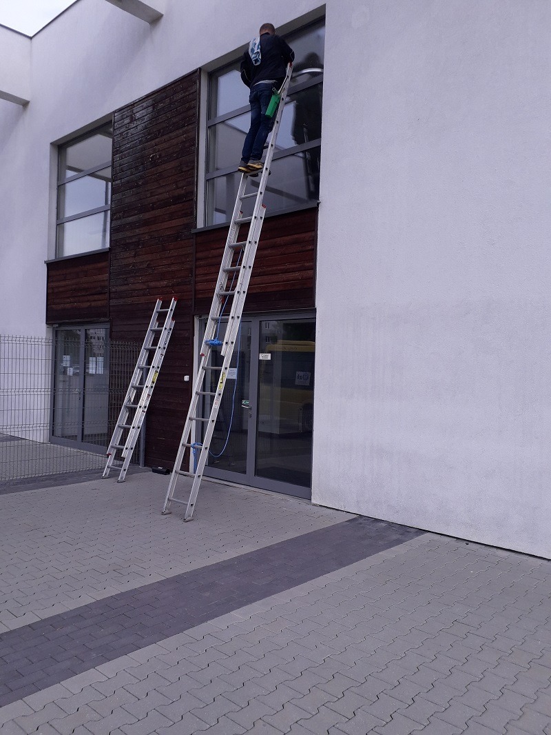 Pracownik w ciemnej kurtce na aluminiowej drabinie czyści okno budynku biurowego; na chodniku stoi druga drabina.