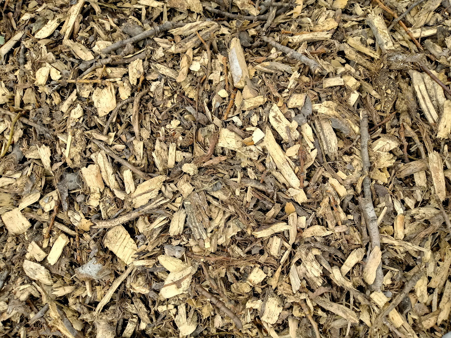 Zrębki drzewne z widocznymi fragmentami kory i gałązek, o zróżnicowanej kolorystyce od jasnego beżu po ciemny brąz, ułożone warstwowo.