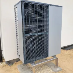 Szara pompa ciepła powietrze-woda na metalowym stelażu, zamontowana na betonowych podstawach obok budynku z jasną elewacją i czarną folią fundamentową.