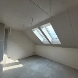 Pokój na poddaszu w trakcie remontu, ściany i sufit wyłożone płytami kartonowo-gipsowymi, widoczne dwa okna dachowe, surowe wykończenie podłogi.