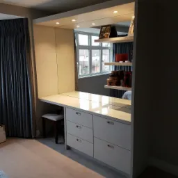 Biała szafa wnękowa z lustrem i biurkiem, oświetlenie punktowe, widok na okno i półki z dekoracjami w odbiciu.