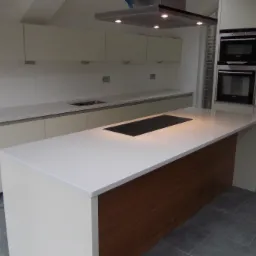 Nowoczesna kuchnia z białą wyspą i drewnianym wykończeniem, płyta indukcyjna na blacie, zabudowane piekarniki i minimalistyczne szafki wiszące.