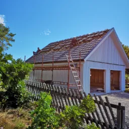 Montaż dachówki na budynku z dwoma bramami garażowymi, widok zza drewnianego płotu i zieleni, pracownicy na rusztowaniu i dachu.