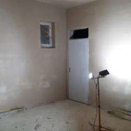 Pok&oacute;j w trakcie prac wykończeniowych: świeżo nałożona warstwa na ścianach i suficie, okno zabezpieczone folią, lampa budowlana oświetla pomieszczenie, podłoga pokryta zabrudzeniami po pracach.