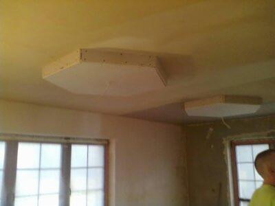 Montaż sufitu podwieszanego z płyt gipsowych w pomieszczeniu z oknami. Widoczne płyty o nietypowym kształcie, przygotowane do dalszego wykończenia.