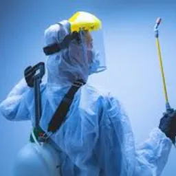 Osoba w białym kombinezonie ochronnym i żółtej osłonie twarzy, z plecakiem-opryskiwaczem, trzymająca lancę opryskową skierowaną w górę, na jednolitym niebieskim tle.