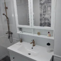 Biała umywalka z szafką i lustrem w łazience z brązową armaturą, widoczna kabina prysznicowa w tle.