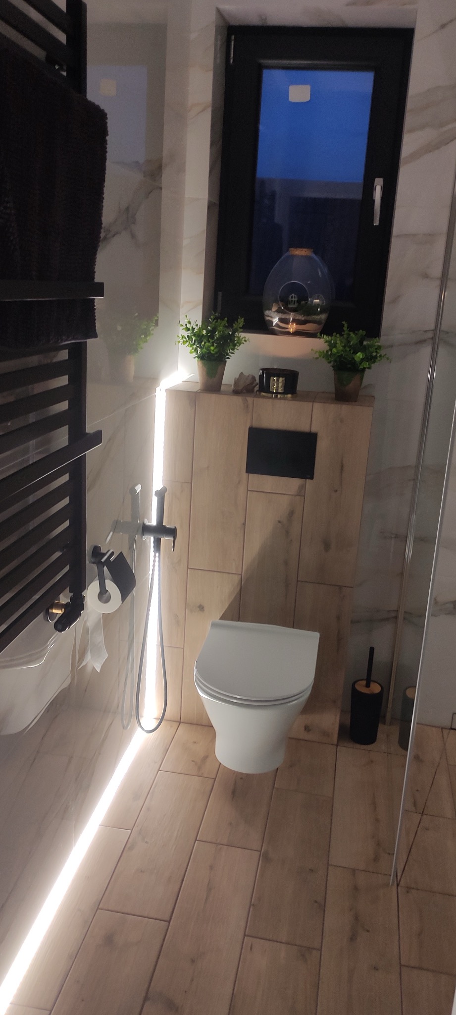 Nowoczesna łazienka z białym sedesem, drewnopodobnymi płytkami na podłodze i ścianie, czarną armaturą i oświetleniem LED wzdłuż ściany, dekoracyjnymi roślinami w doniczkach na zabudowie geberit...