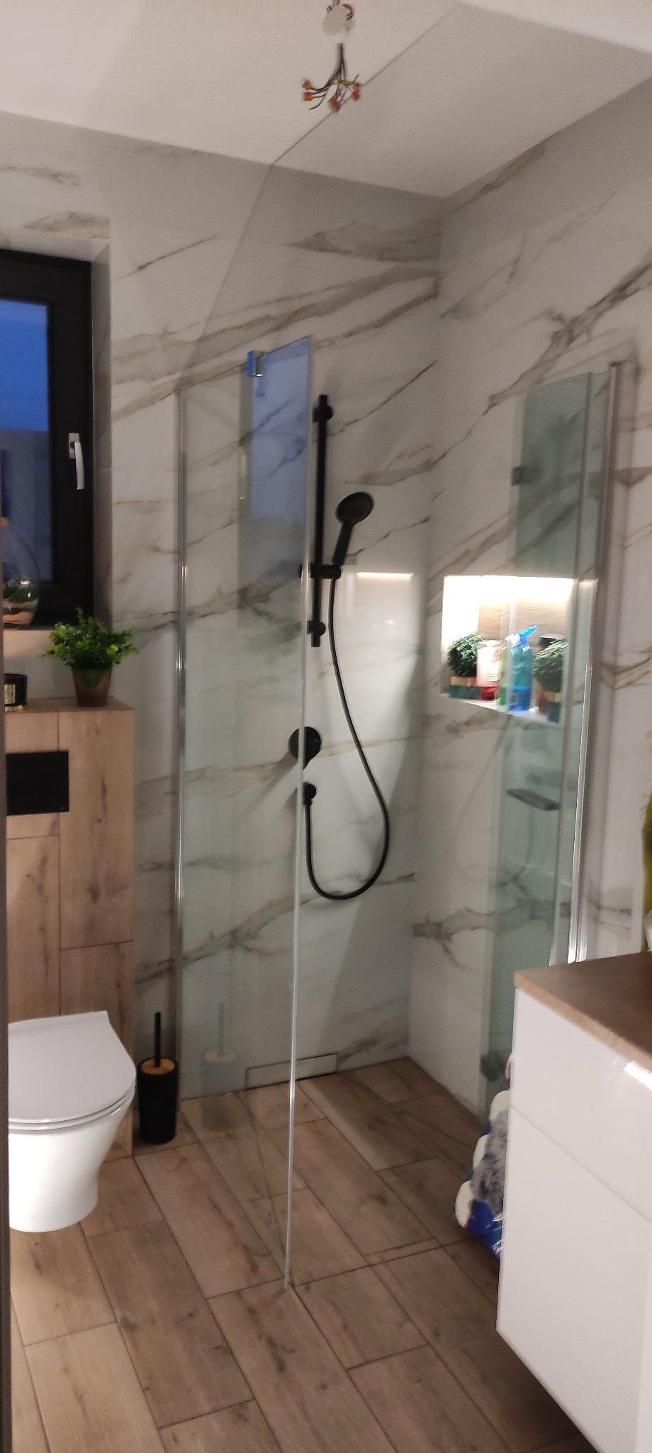 Nowoczesna łazienka z kabiną prysznicową typu walk-in, wykończona płytkami imitującymi marmur i drewno, widoczna toaleta i czarne akcesoria.