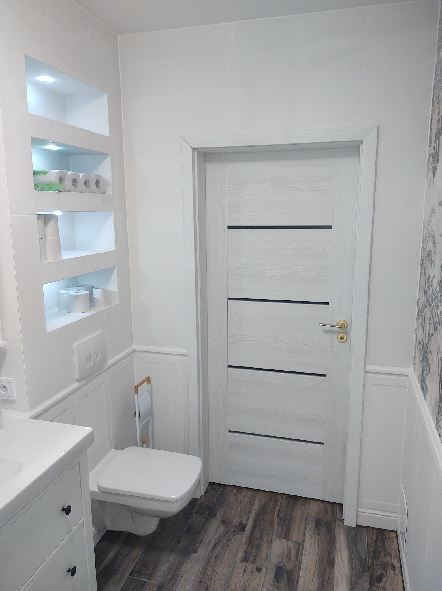 Biała łazienka z drewnopodobną podłogą, białą zabudową, toaletą podwieszaną i drzwiami z poziomymi, czarnymi wstawkami oraz wnęką z oświetleniem i zapasem papieru toaletowego.