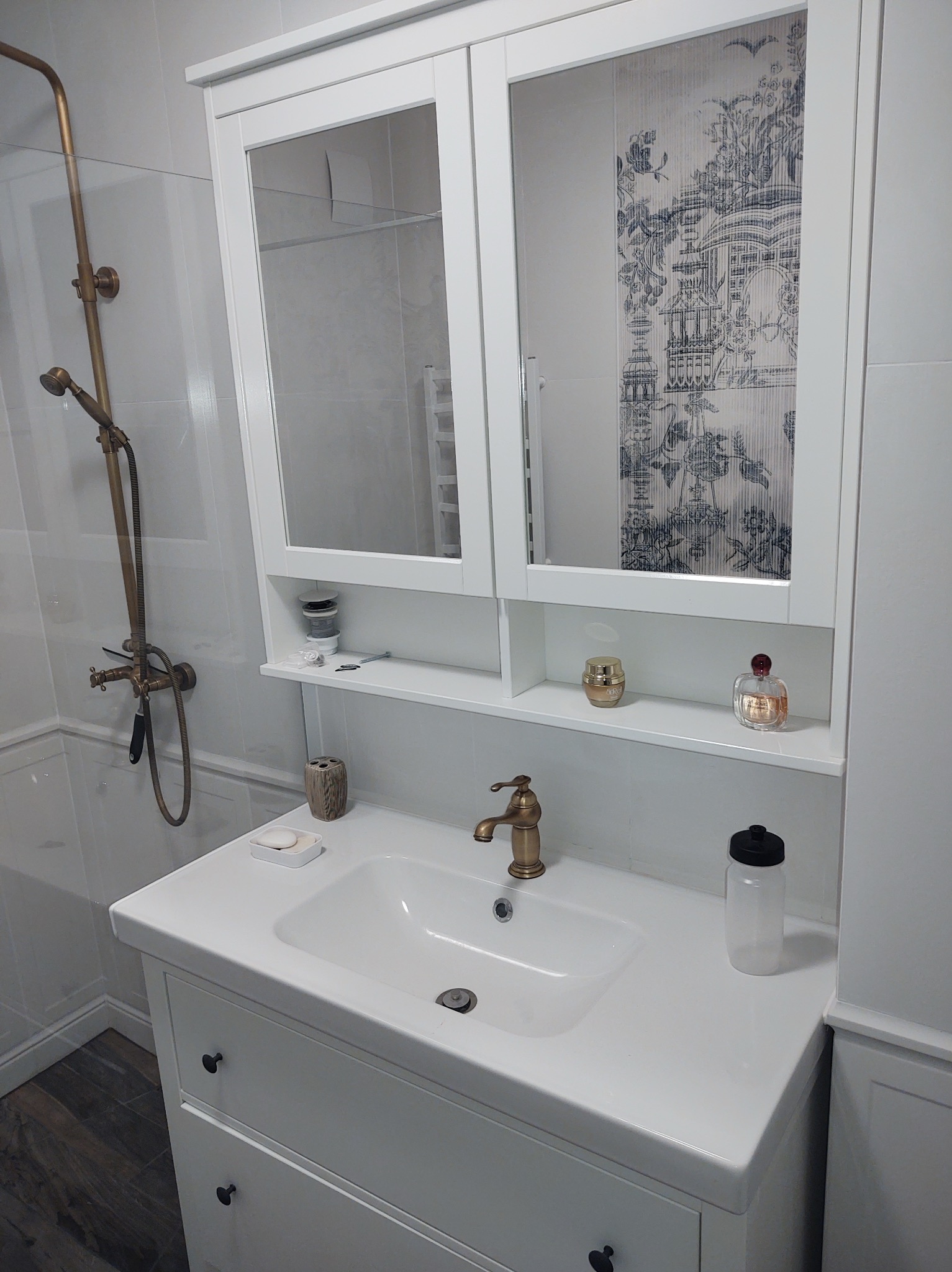 Biała umywalka z szafką i lustrem w łazience z brązową armaturą, widoczna kabina prysznicowa w tle.