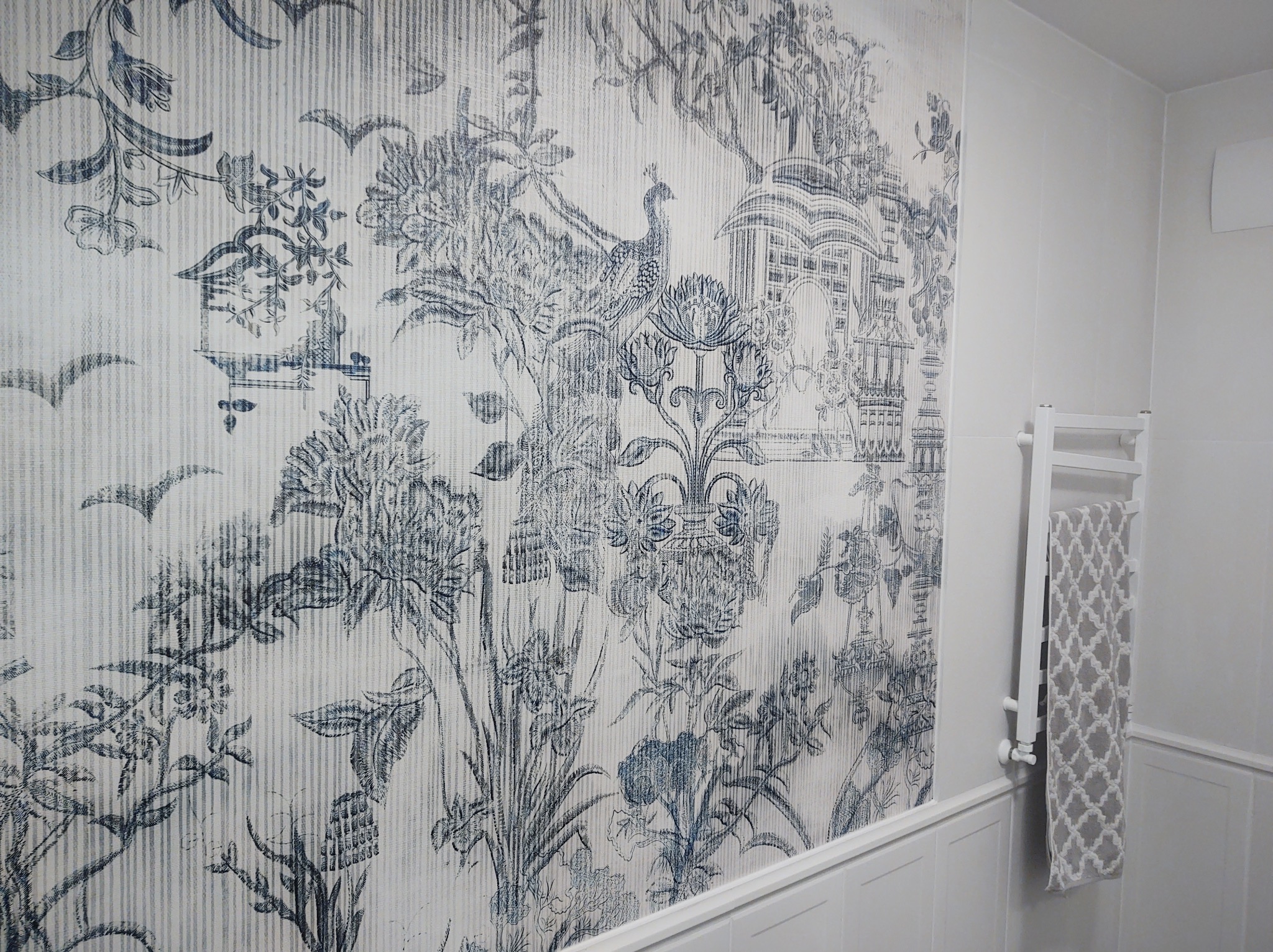 Ściana w łazience pokryta tapetą w stylu toile de jouy z motywem pawia i roślinności, biała grzejnik drabinkowy z szarą ręcznikiem w romby.
