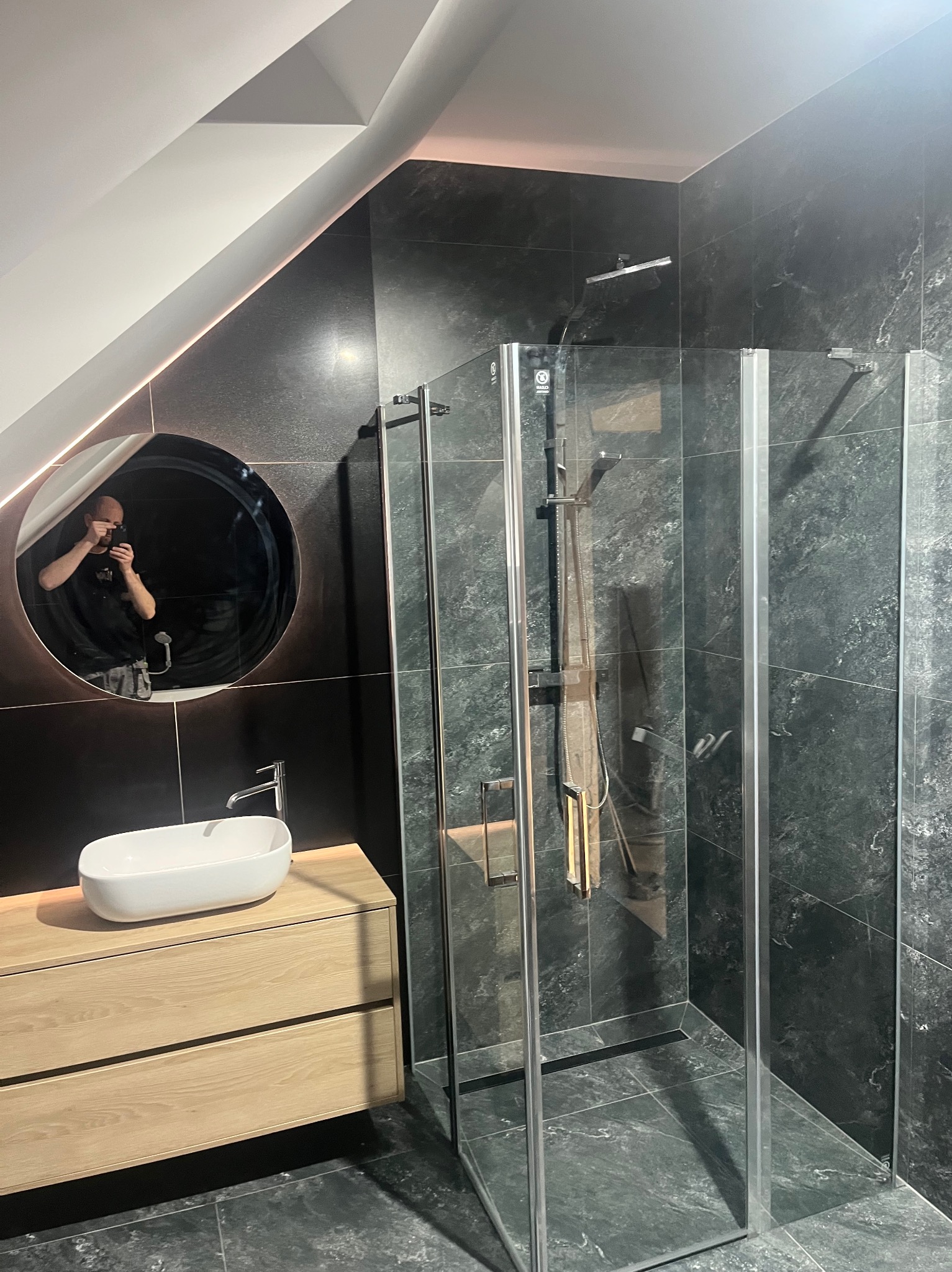 Nowoczesna łazienka z kabiną prysznicową typu walk-in, umywalką nablatową na drewnianej szafce i okrągłym lustrem z podświetleniem LED. Czarno-szare płytki imitujące kamień.