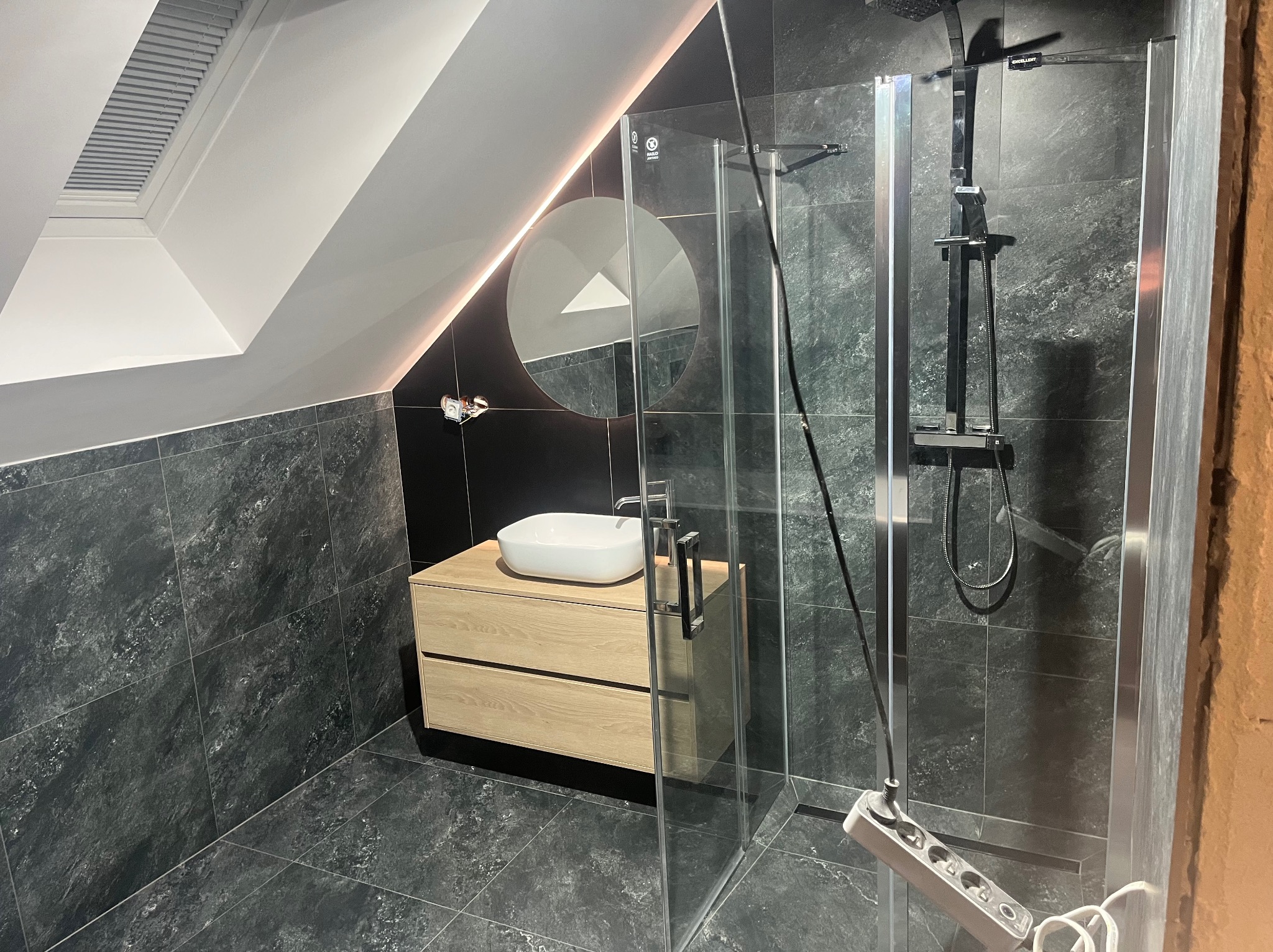 Nowoczesna łazienka na poddaszu z czarnymi płytkami, drewnianą szafką pod umywalką i szklaną kabiną prysznicową. Okrągłe lustro z podświetleniem LED.