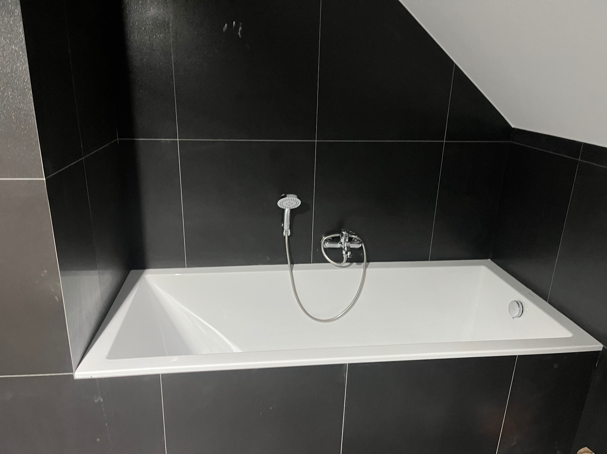 Nowoczesna łazienka z czarnymi płytkami i białą wanną pod skosem dachu. Minimalistyczny design, chromowana armatura, czyste linie i ostre kąty.