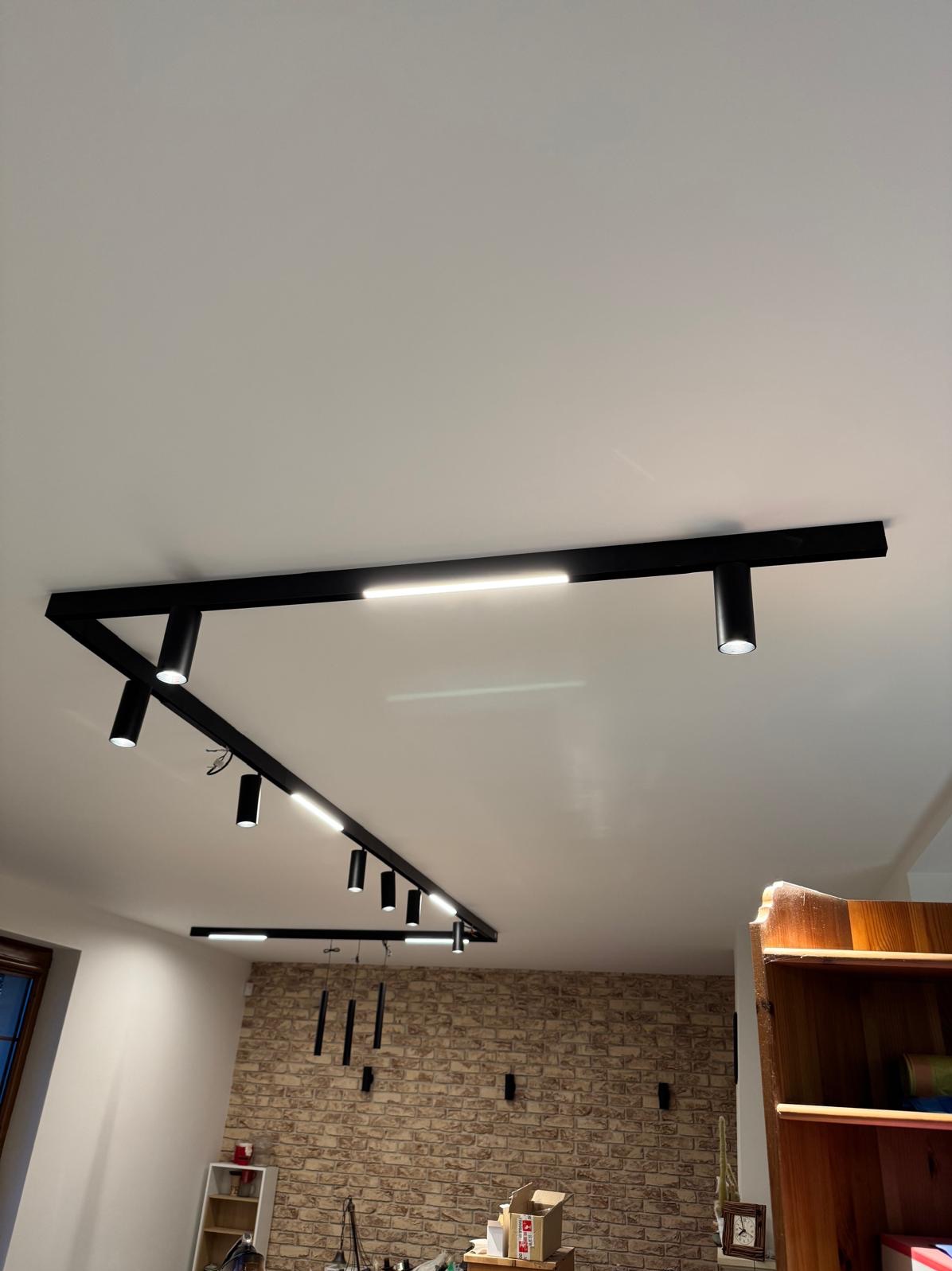 Nowoczesne oświetlenie sufitowe z czarnymi szynami i reflektorami LED, skierowane w dół, na tle ściany z cegły i jasnego sufitu.