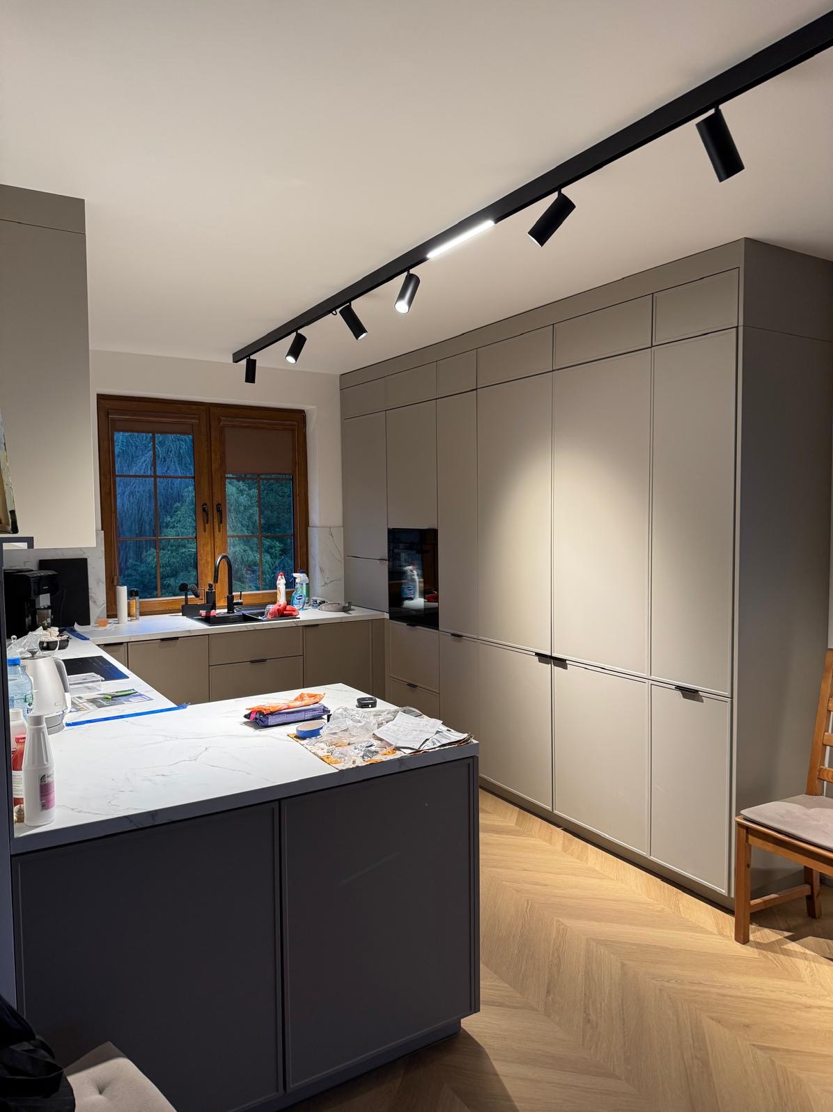 Nowoczesna kuchnia w odcieniach szarości z wyspą i oświetleniem szynowym. Parkiet ułożony w jodełkę. Widok na okno z zielenią na zewnątrz.