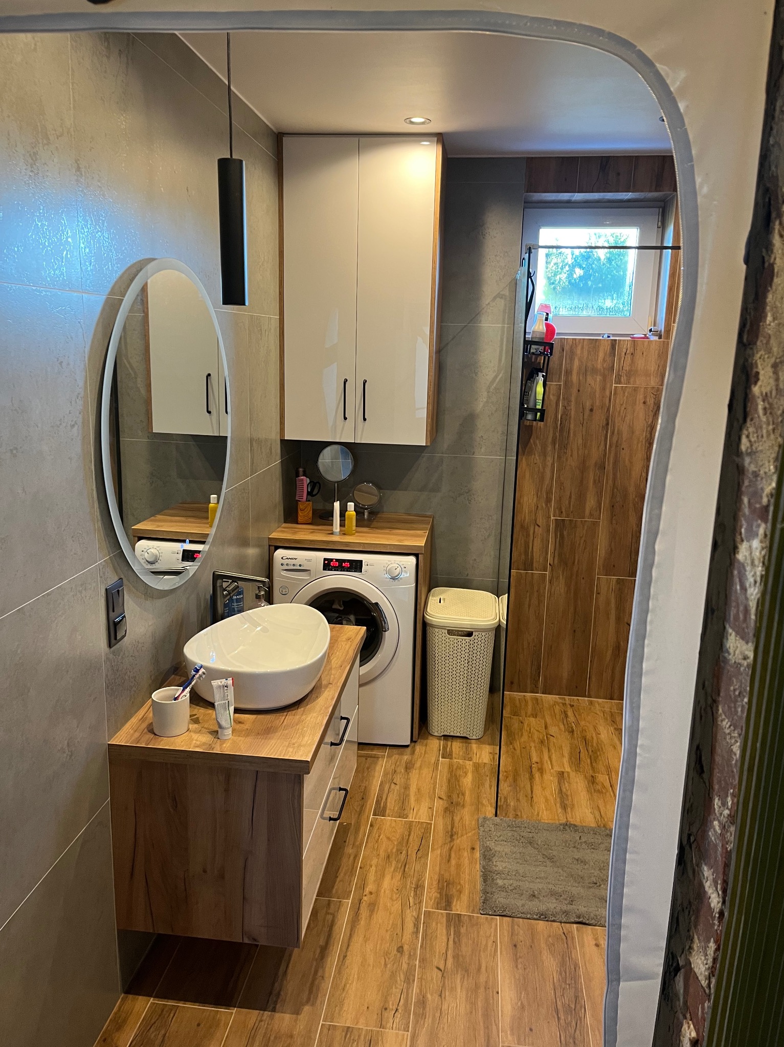 Nowoczesna łazienka z owalnym lustrem, drewnianą szafką pod umywalką i pralką. Szare płytki na ścianach i podłodze imitujące drewno. Minimalistyczna lampa wisząca.