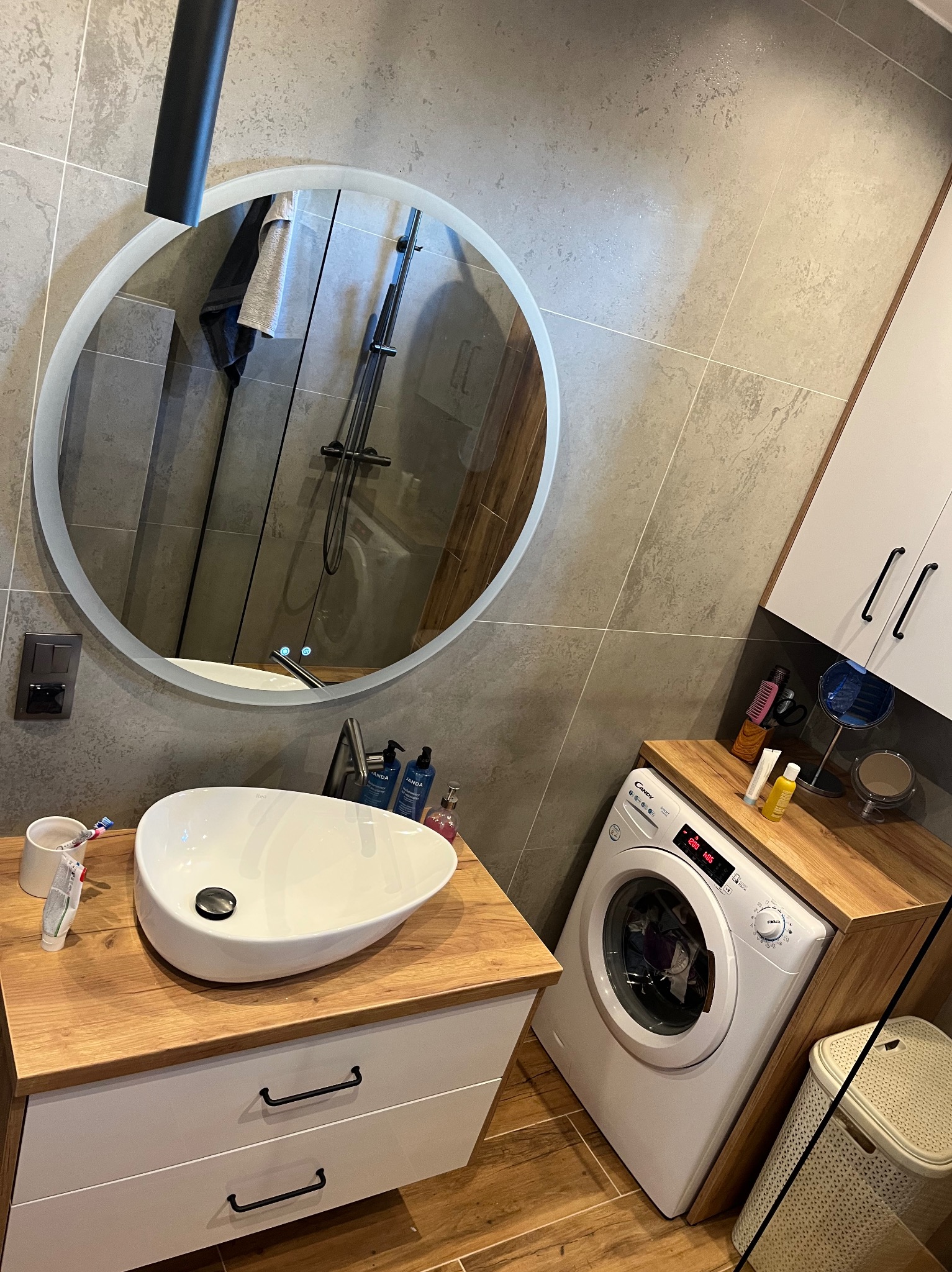 Nowoczesna łazienka z okrągłym lustrem LED, umywalką nablatową i pralką pod drewnianym blatem. Szare płytki i białe szafki tworzą spójną całość.